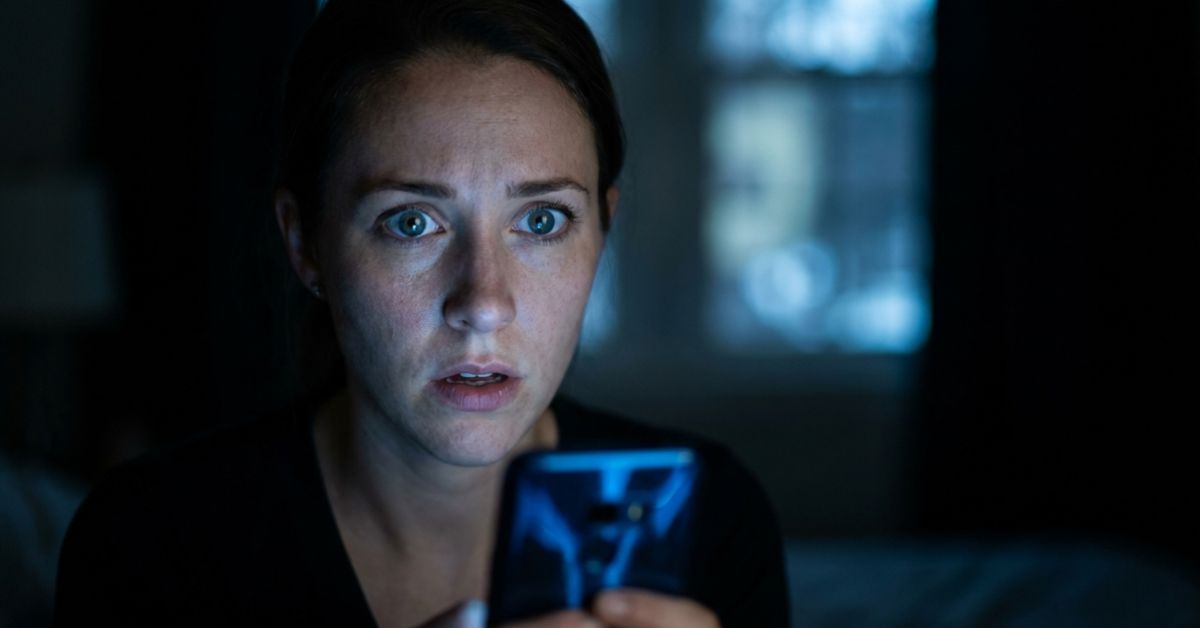 A woman looks at her smartphone with a shocked and alarmed expression in a dark room. Her eyes are wide with worry, and her mouth is slightly open. The blue light from the phone screen illuminates her face against the dark background.