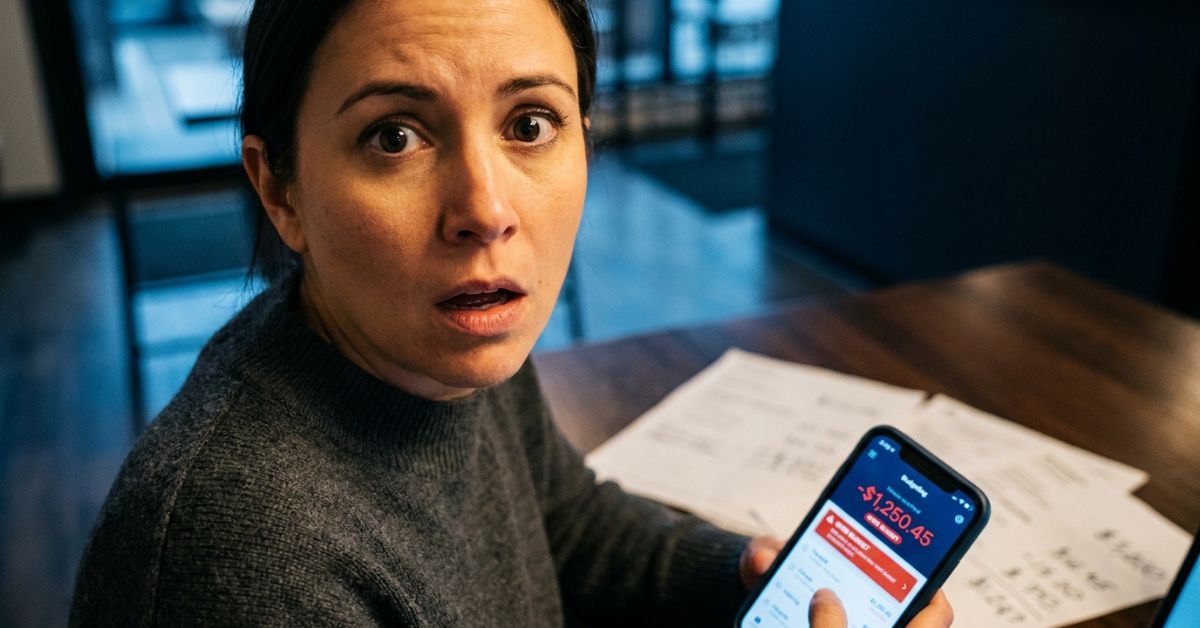 A woman with a stunned and concerned expression looks directly at the camera while holding a smartphone. The phone screen displays a budgeting app with a large negative balance of -$1,250.45 and a red "Over Budget" alert. She is sitting at a wooden table with several papers scattered behind the phone, suggesting she is reviewing her finances.