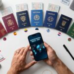A top-down view of a white desk where a person holds a smartphone displaying a "Digital Nomad Visa Options - 2026" app. Surrounding the phone are passports from various countries including Thailand, Spain, Portugal, and Greece, along with boarding passes, a compass, and sticky notes detailing income and savings requirements.