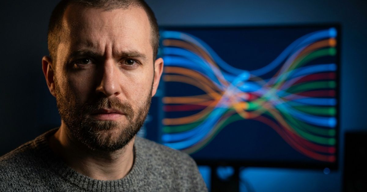 A close-up portrait of a man with a short beard and a concerned expression, looking directly at the camera. He is wearing a grey knit sweater. Behind him, a large computer monitor glows in a dark room, displaying an intricate data visualization with many colorful, intertwined curved lines.